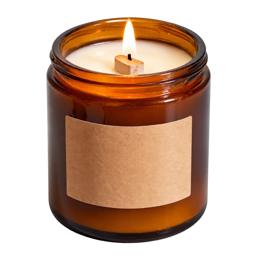 An amber glass jar candle with a blank kraft paper label and a lit wooden wick, centred on a pure white background