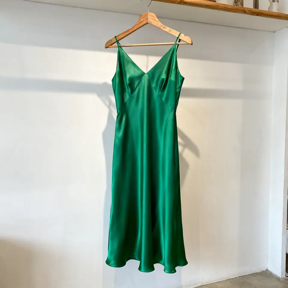 Vestido midi de satén verde esmeralda en una percha de madera contra la pared blanca de una boutique